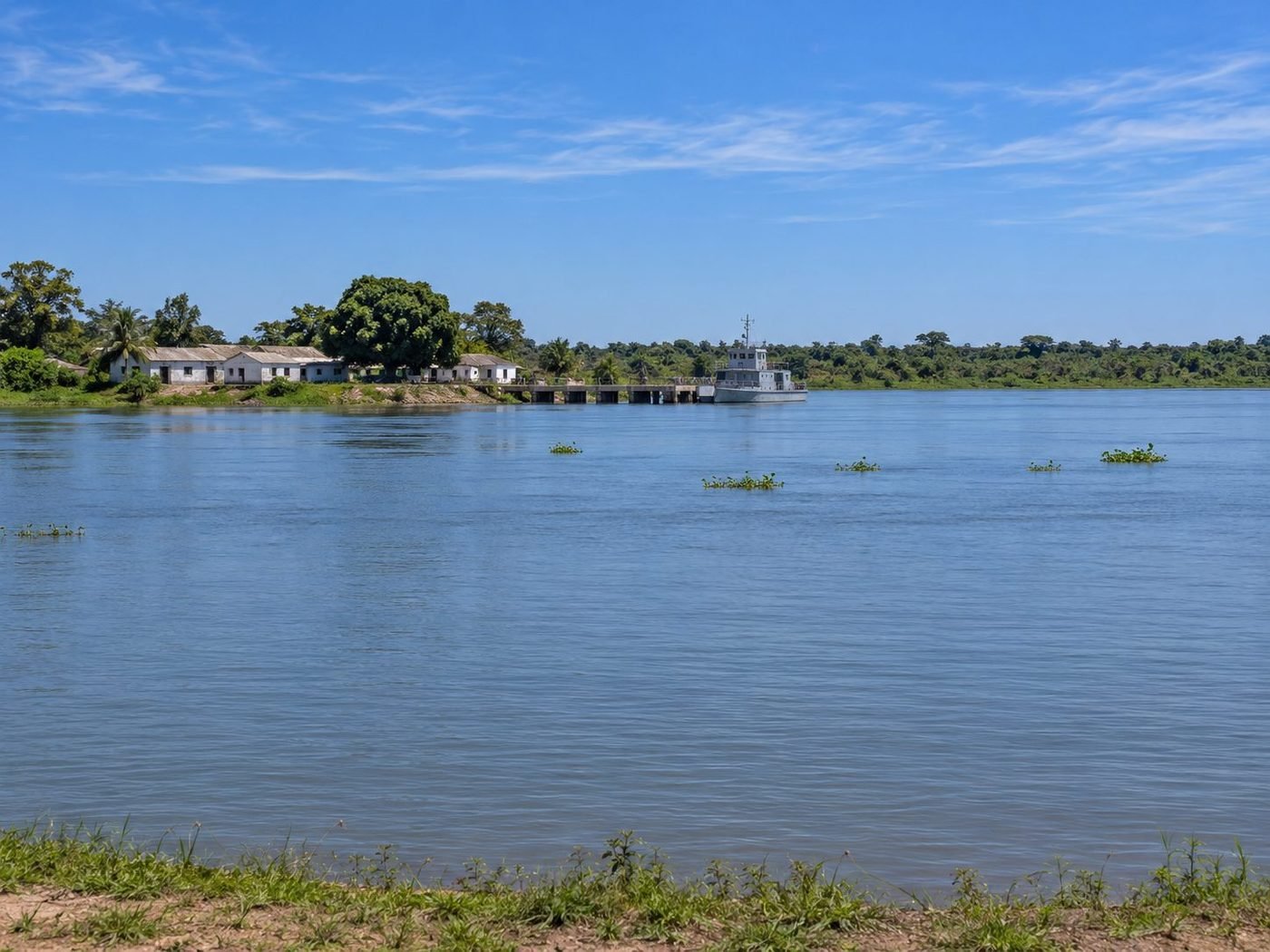Navio cinza atracado no píer de um rio pantaneiro, com casas brancas na margem, paisagem como a de Ladário (MS)